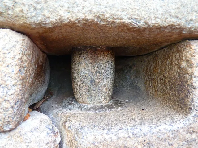 Un bâtiment en ruine avec seulement une partie de la toiture en tuile, les murs sont en pierre et on y voit une fenêtre aux bords arrondis. Au fond il y a un portail en tôle.