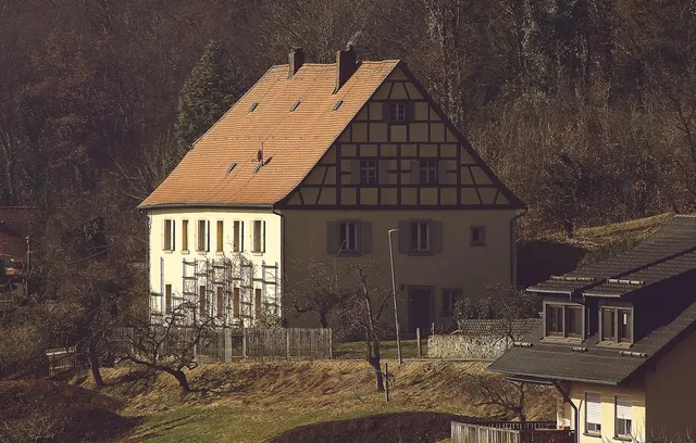 Vue de la bâtisse principale, avec un toit en tuile, des murs en béton et des fenêtres avec des volets fermés qui sont en bois.