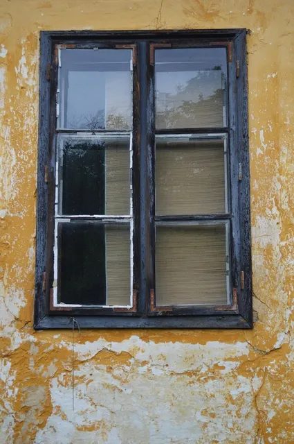 Bâtiment principal de la parcelle, il y a 6 fenêtres avec volets fermés et deux portes, elles aussi condamnées. La pelouse est verte et il y a un côté avec plusieurs arbres à gauche.