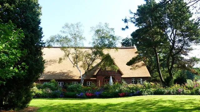 La maison principale vue de loin avec sa toiture en tuile et ses murs en béton. On y voit également les différents arbres derrière et sur les côtés.